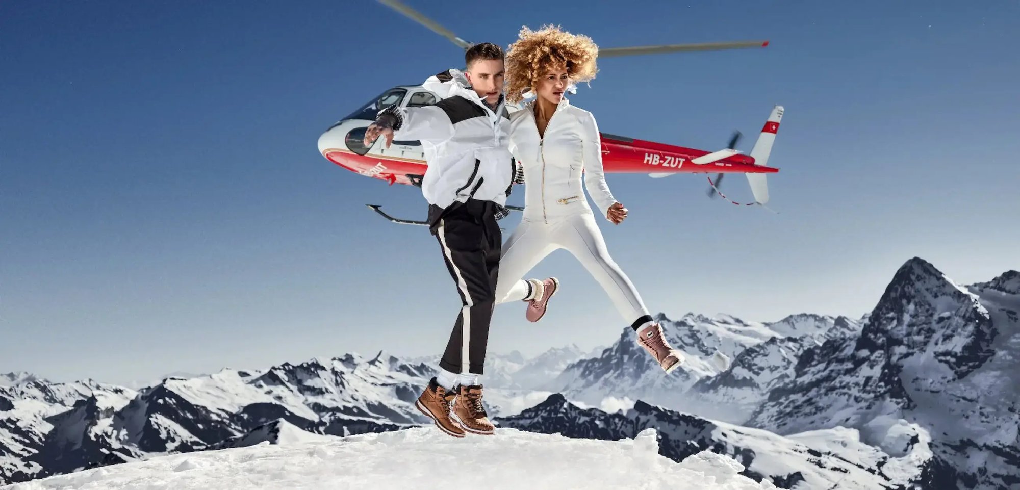 In einer verschneiten Berglandschaft springen zwei Personen in die Luft. Hinter ihnen ist ein Helikopter zu sehen.
