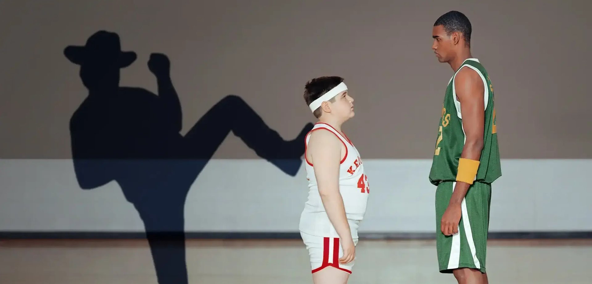 Ein Junge steht einem viel grösseren Basketball-Spieler gegenüber. Hinter dem Jungen ist der Schatten eines Mannes zu sehen, der dem Jungen scheinbar einen Kick in den Rücken gibt.