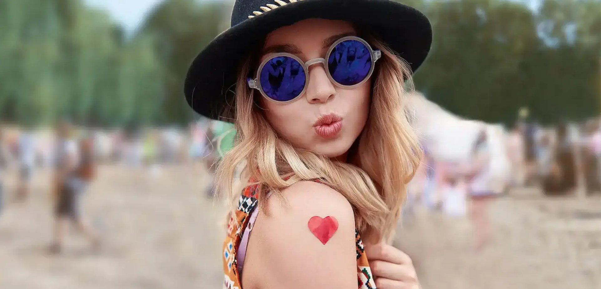 Junge Frau an einem Festival mit Sonnenbrille und Hut. Sie trägt einen roten Herz-Sticker auf dem Oberarm.