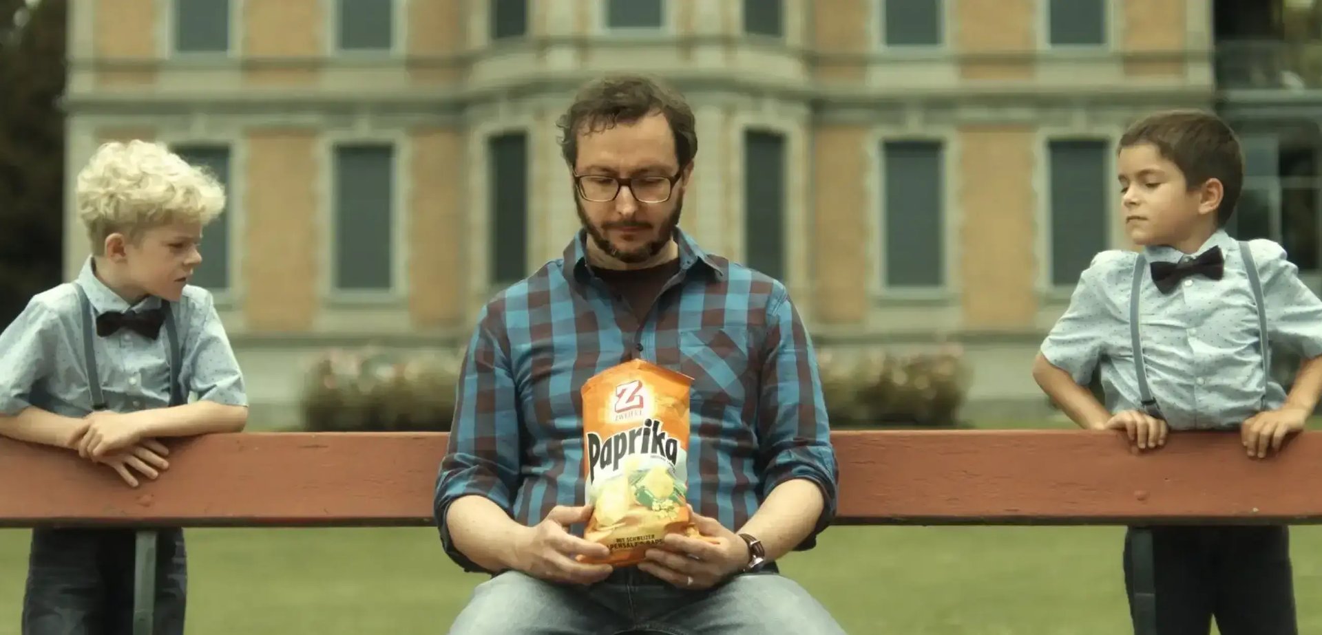 Ein Mann sitzt auf einer Bank und blickt in eine geöffnete Zweifel-Chips-Packung. Rechts und links von ihm steht jeweils ein Junge.