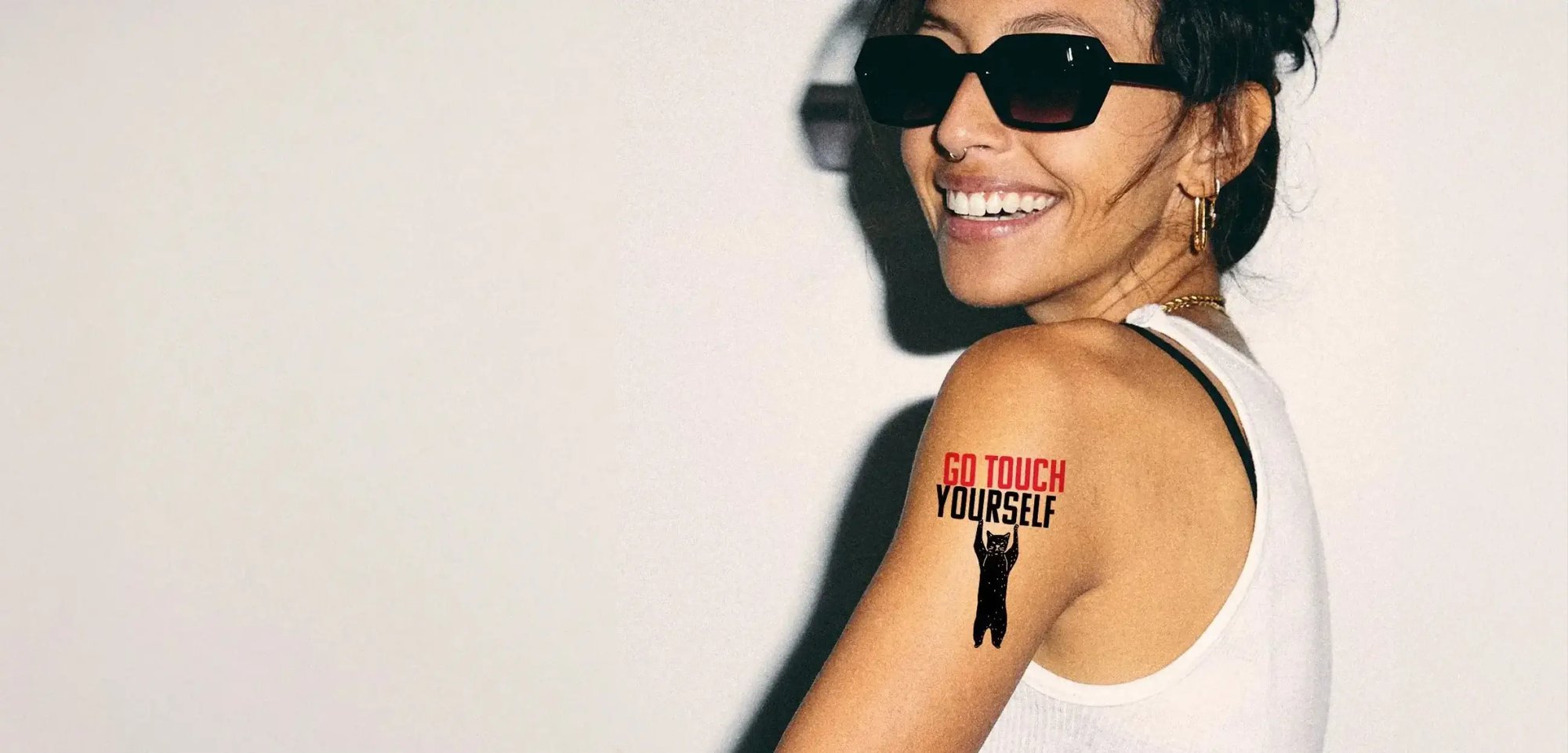 Eine Frau mit Sonnenbrille trägt auf dem Oberarm ein temporäres Tattoo auf welchem "Go Touch Yourself" geschrieben steht.