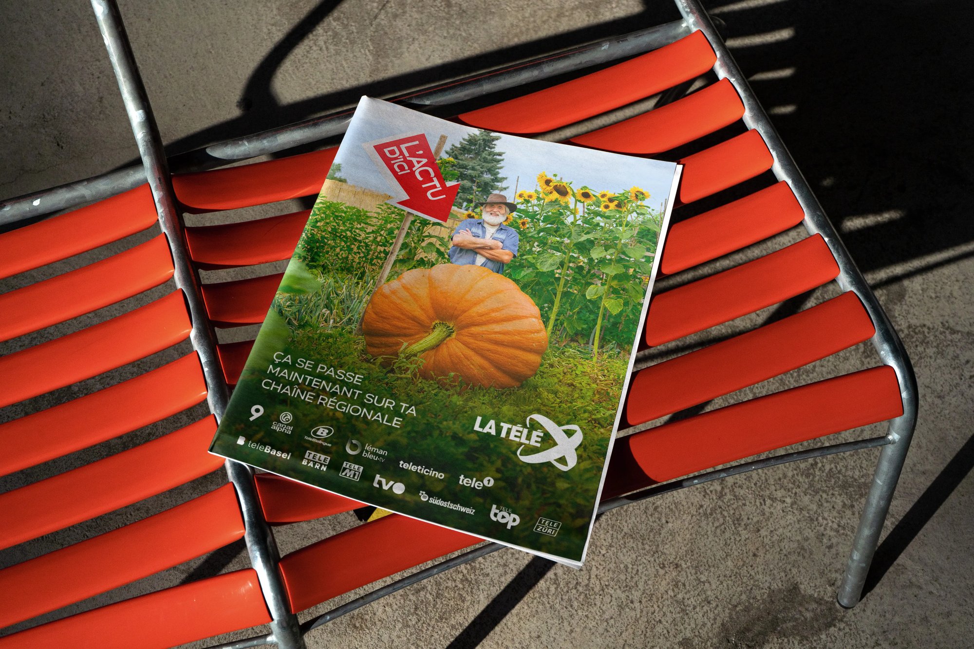 Auf einer Bank liegt ein Magazin auf welchem das Bild eines Mannes in einem Schrebergarten zu sehen ist. Der Mann steht hinter einem grossen Kürbis. Ein Schild in Form eines Pfeils zeigt auf den Kürbis. Auf dem Pfeil steht "News von hier".