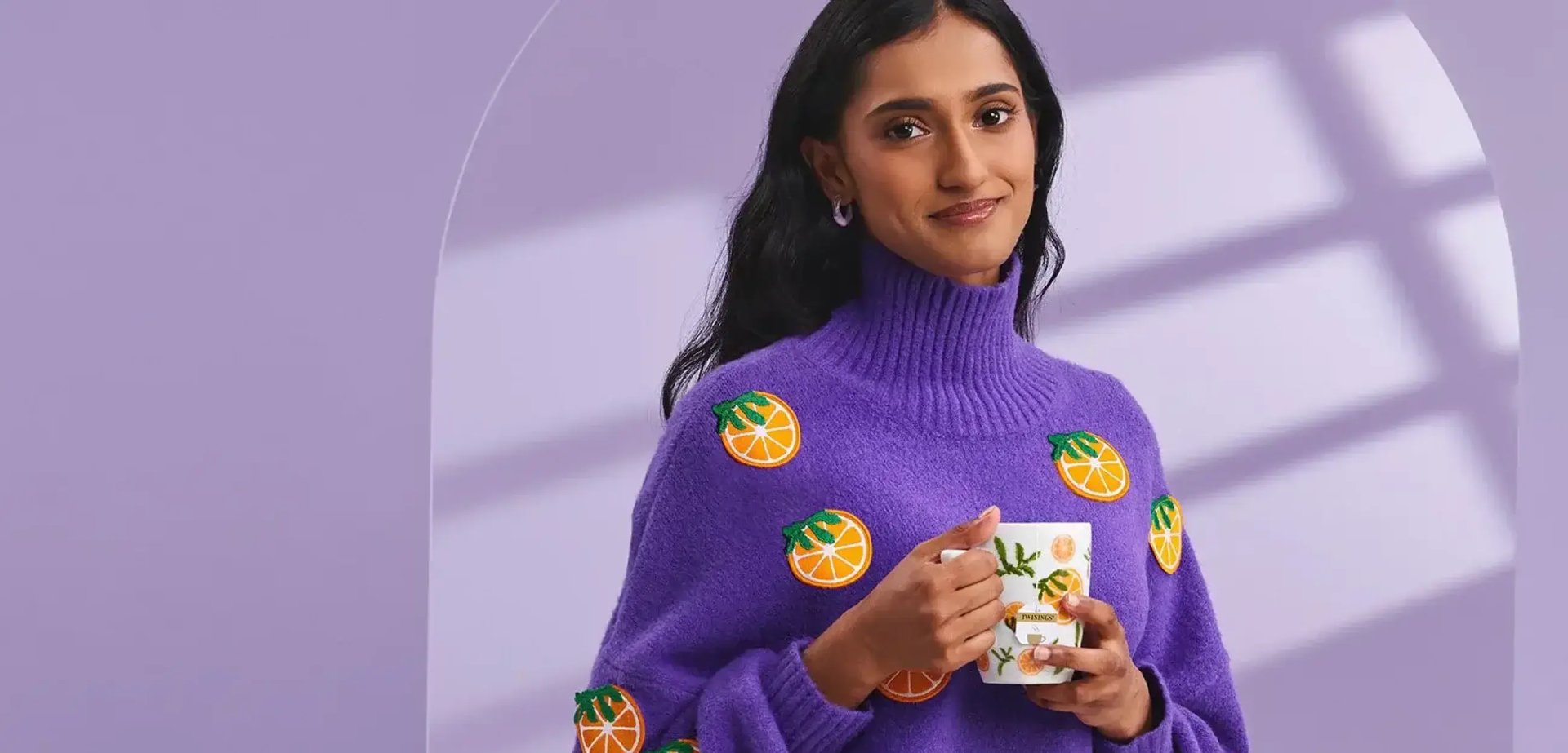 Eine Frau trägt einen Pullover, auf dem Orangen aus Stoff aufgenäht sind. Sie trinkt aus einer Teetasse, auf welcher ebenfalls Orangen zu sehen sind.