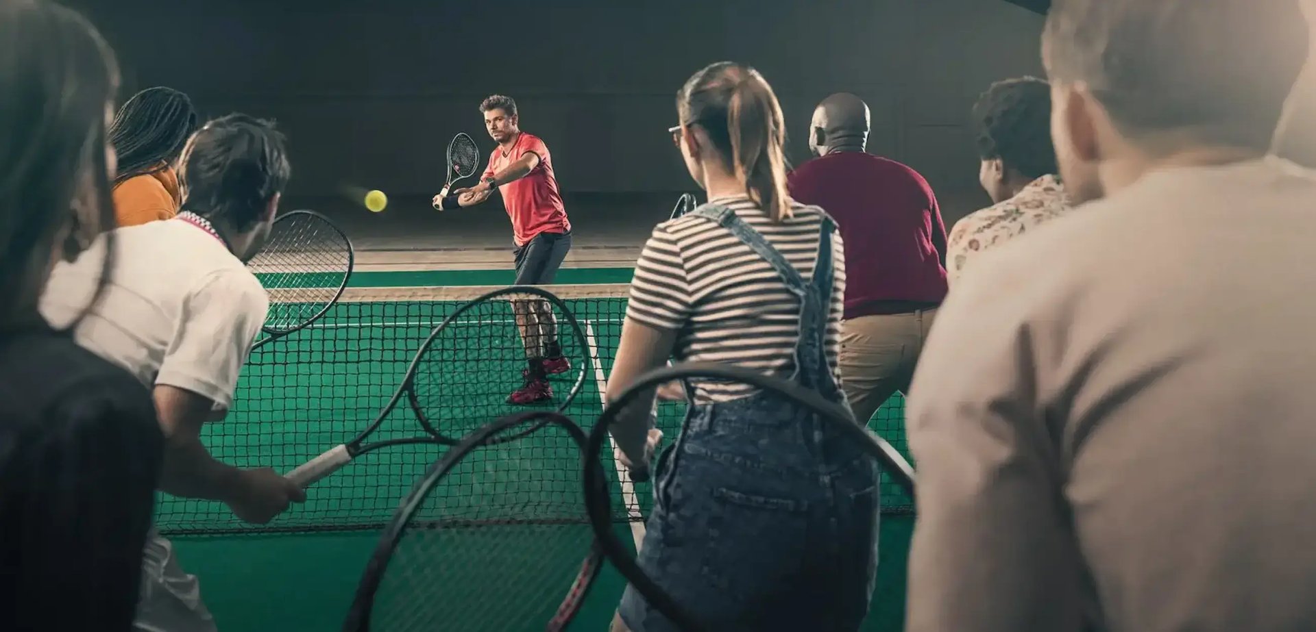 Tennnis-Star Stan Wawrinka steht auf einer Seite eines Tennis-Platzes und macht einen Aufschlag. Auf der anderen Seite des Platzes stehen viele Menschen mit Tennisschlägern bereit.