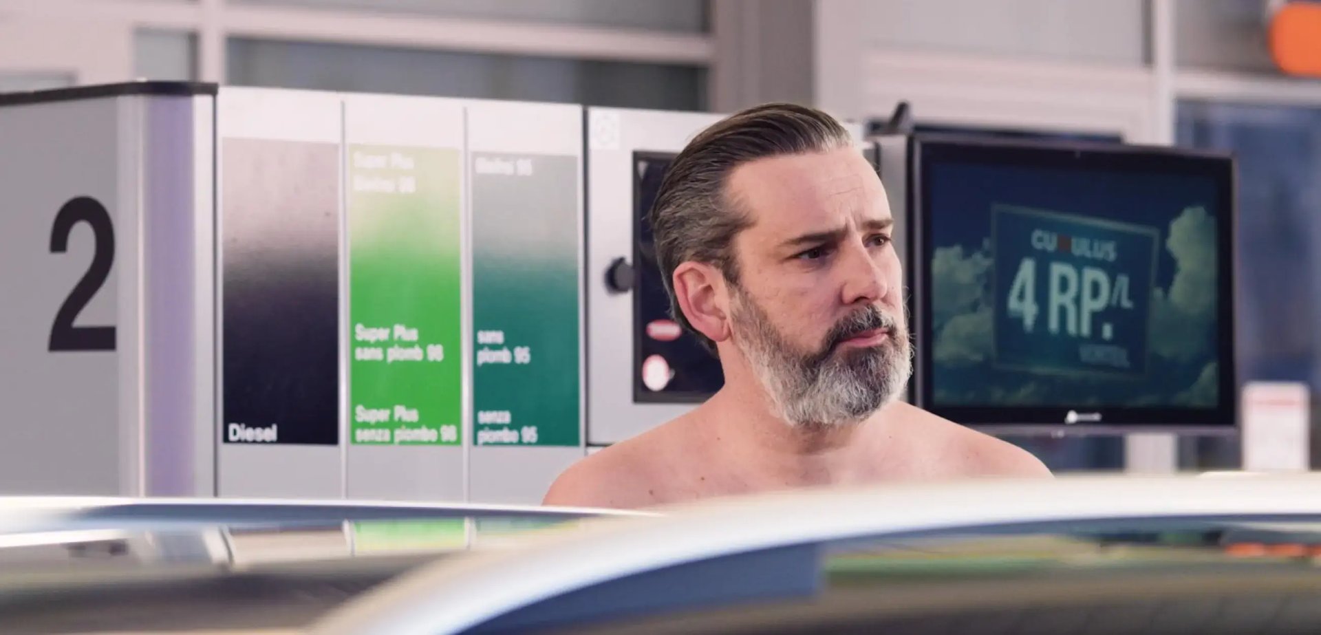 Ein Mann mit freiem Oberkörper steht an einer Tankstelle neben der Zapfsäule und einem Auto.