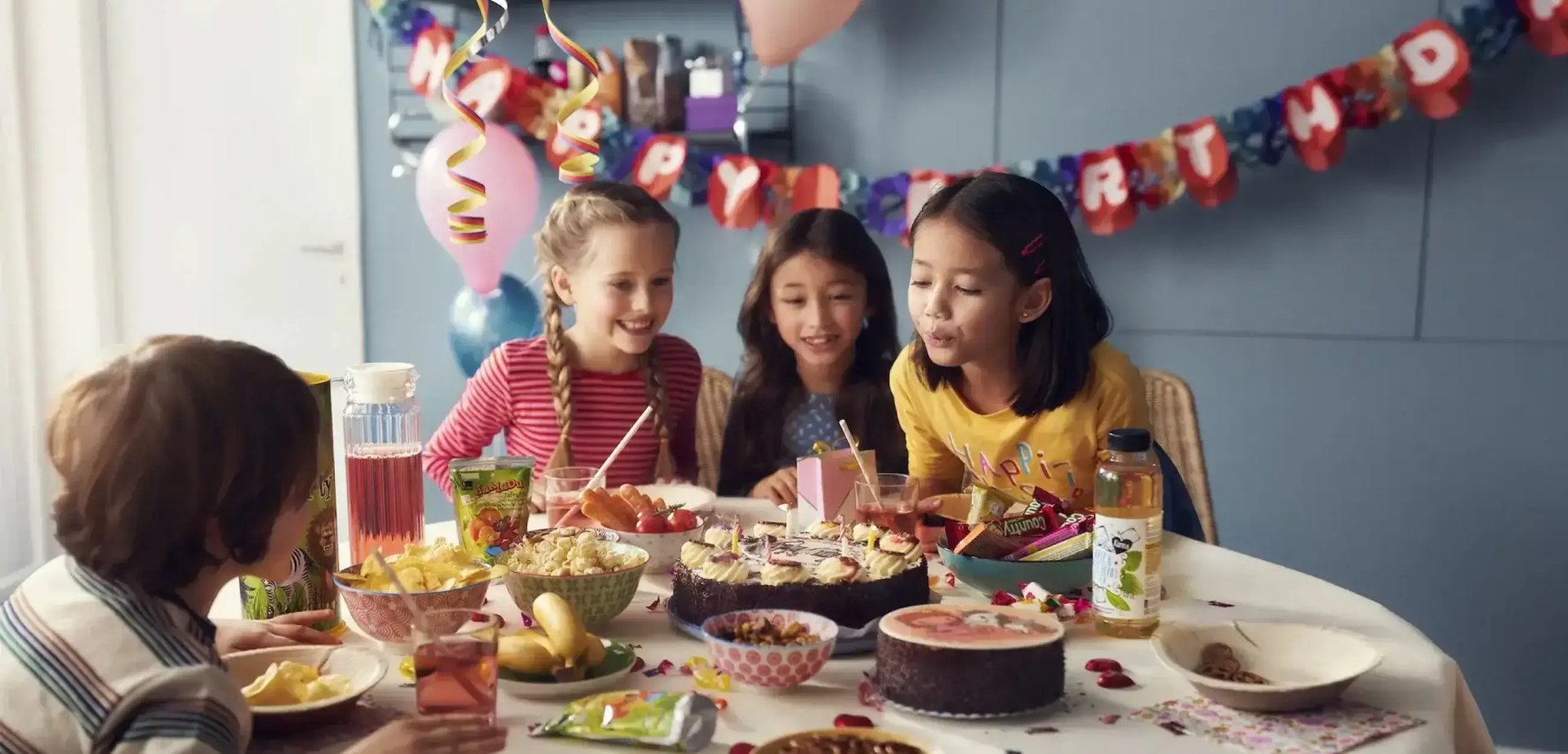 Drei Kinder an einem gedeckten Tisch an einem Kindergeburtstag. Ein Mädchen bläst Kerzen auf einem Kuchen aus.