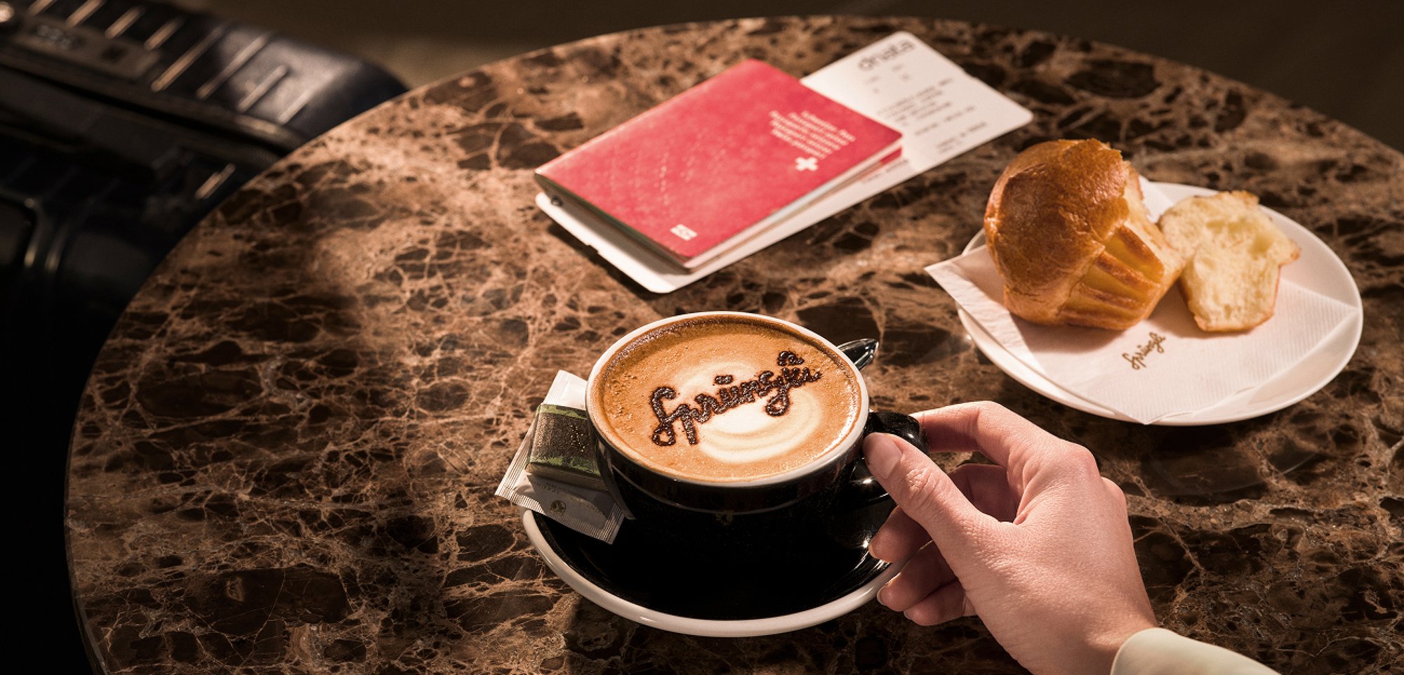 Auf einem Marmortisch stehen ein Cappuccino, ein Muffin und ein Schweizer Reisepass. Eine Hand greift nach der Kaffee-Tasse. Neben dem Tisch steht ein Reisekoffer.