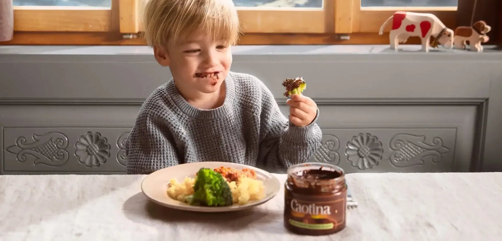 Ein Kind mit schokoladenverschmierten Gesicht sitzt an einem Esstisch und hat ein Stück Brokkoli in der Hand, welches in Caotina Creme getaucht wurde.